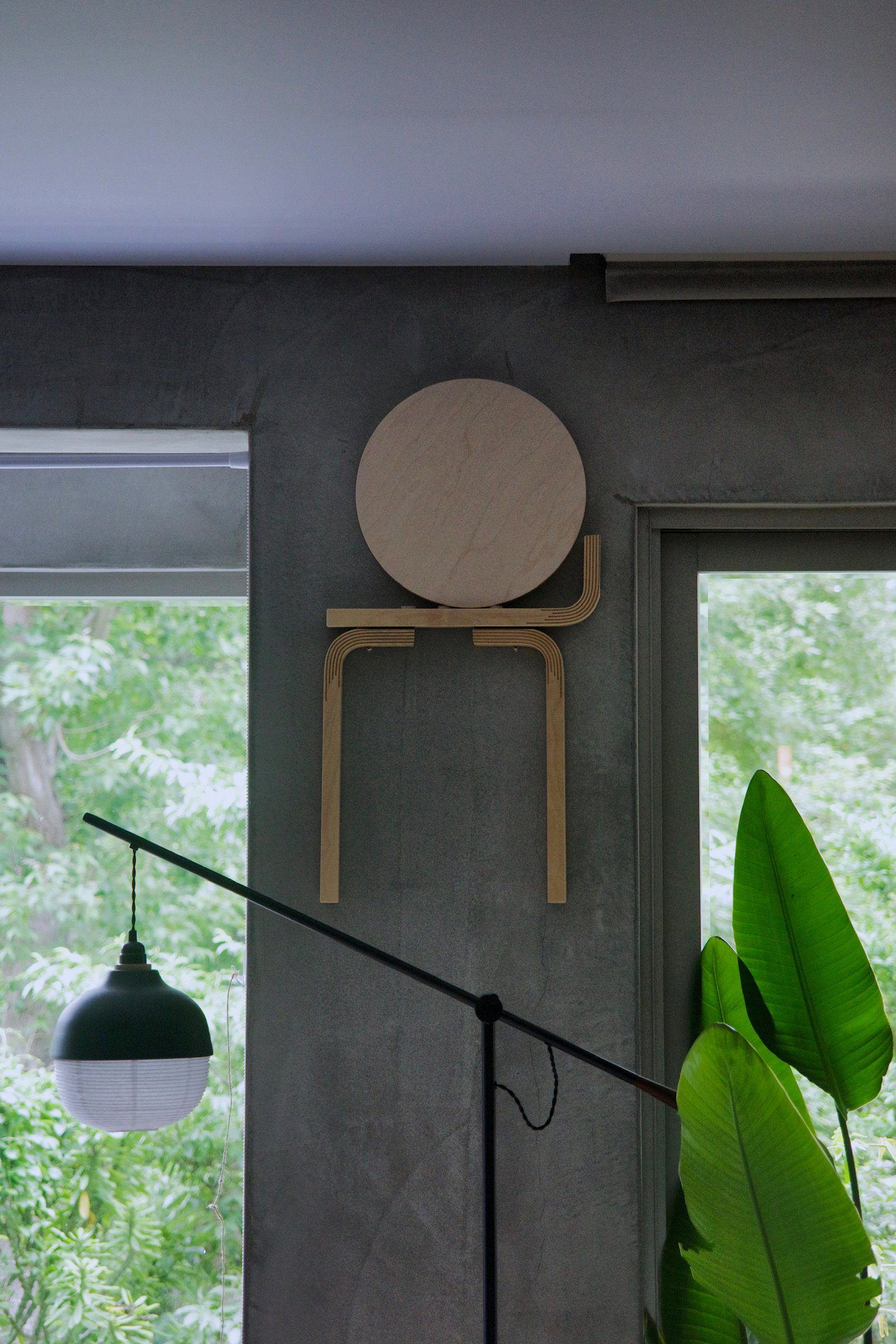 Artek Stool 60 the Kontrasti edition disassembled and hung on a concrete wall as art. Greenery outside the window.