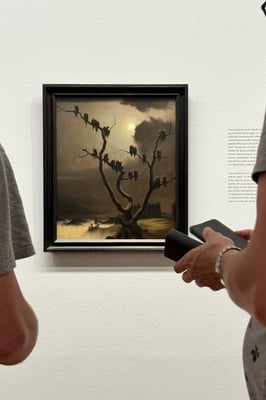 A painting on the wall in a museum with passer by. Paining has an erring feeling with a dying tree with what seems like crowd perching but are actually skully figures in black robes.
