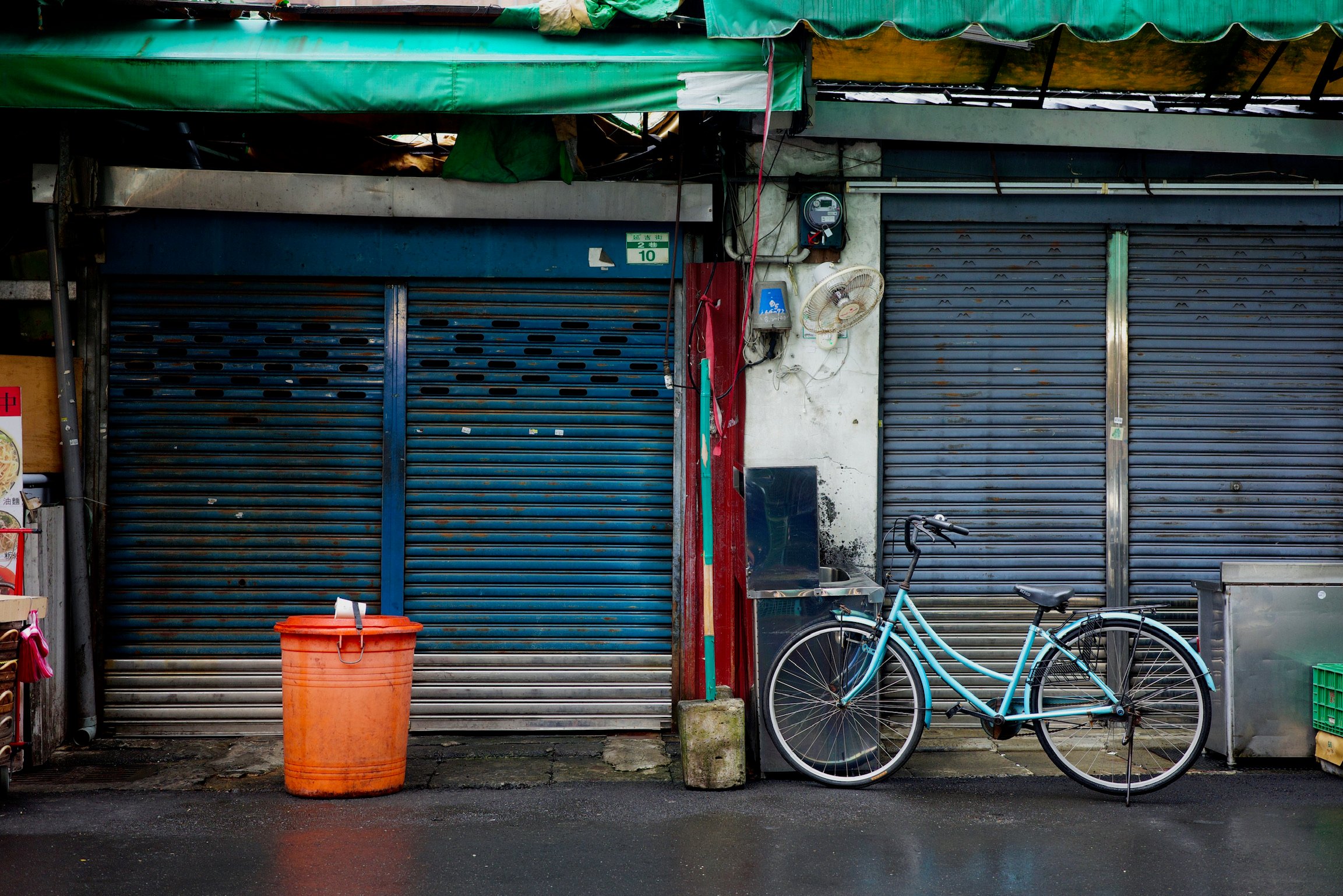 Rainy Taipei, blue roll-up door, orange bin, sky blue bike, wet asphalt.