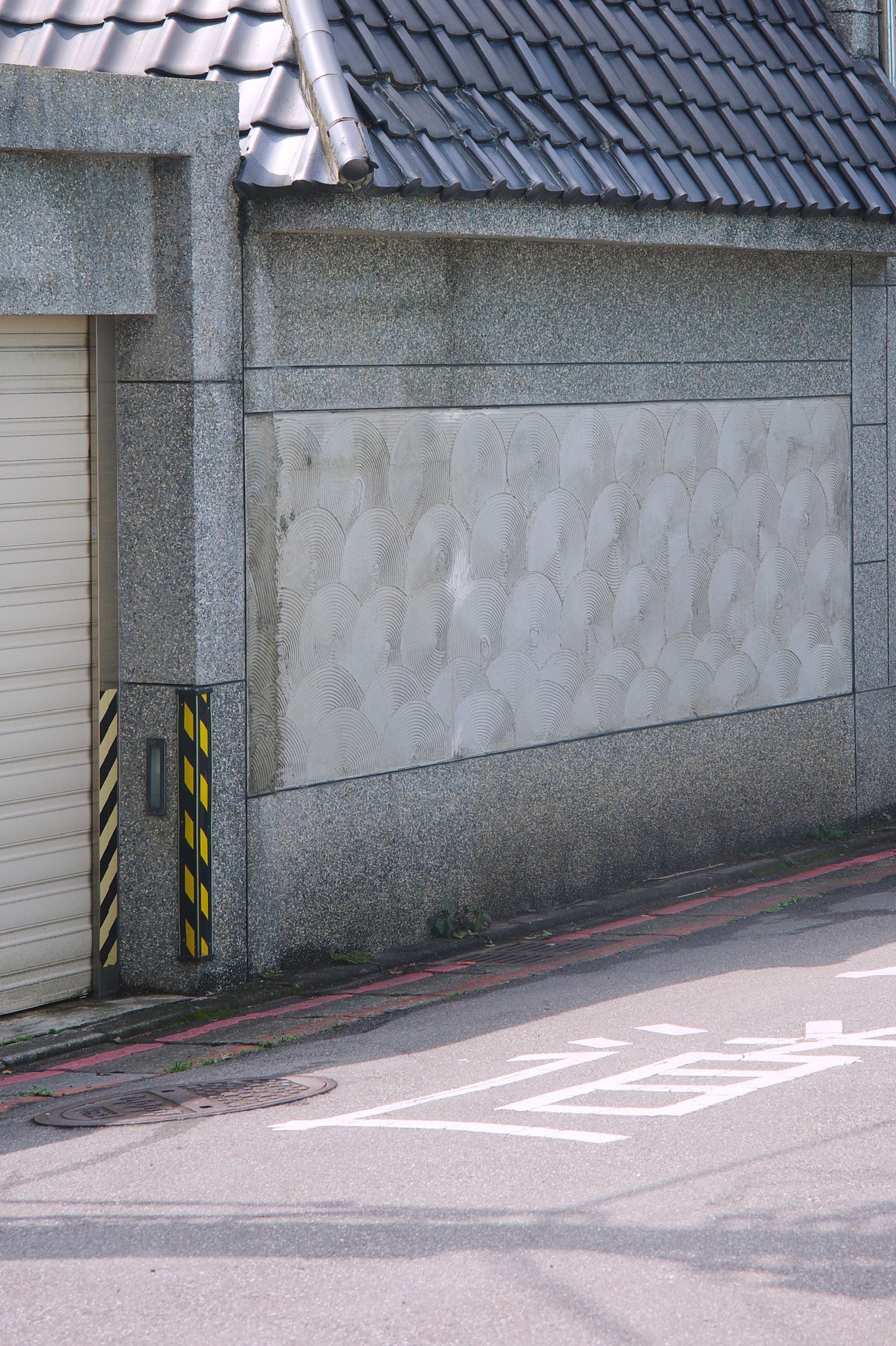 Sunny day, front of a garage door. The wall beside has repeating round brush patterns and dark blue paneling roof.