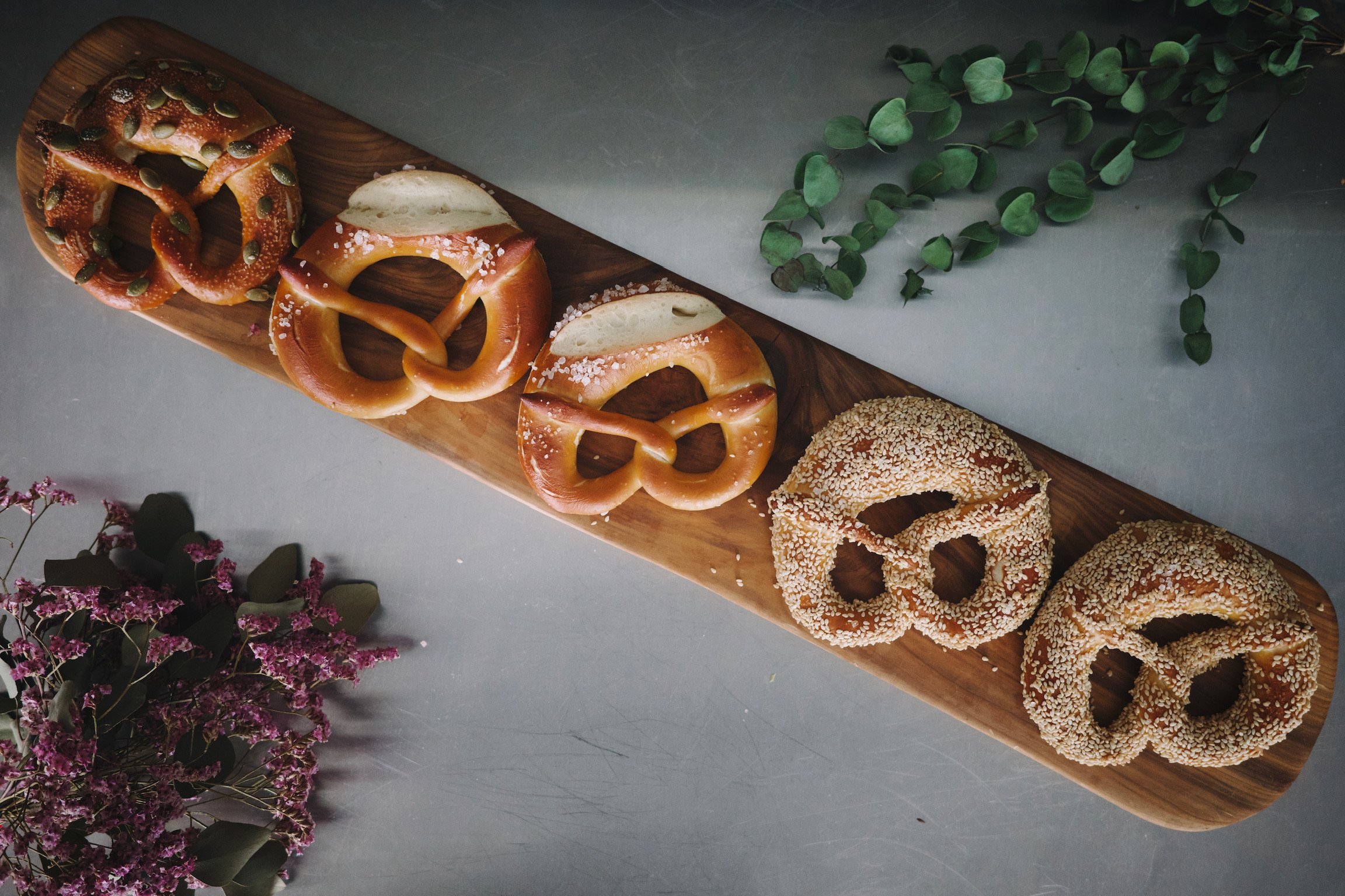 5 pretzels on a wooden plank on top of a stainless steel counter with dried flower bouquets as decorations.