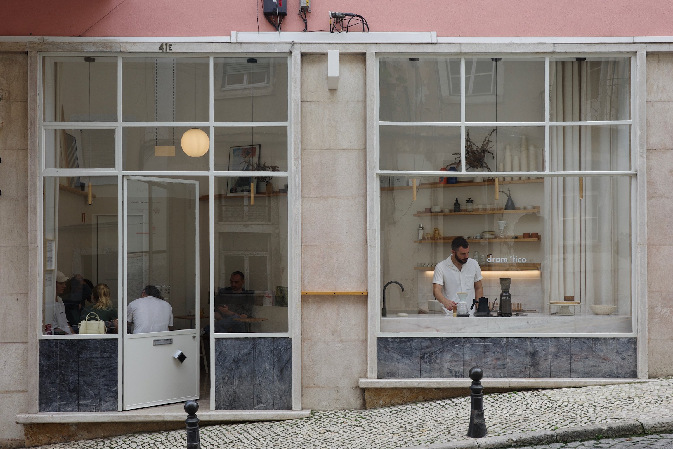 A tiny cafe on a slope with pink external walls and white floor to ceiling window frames and blocks of grey panels.