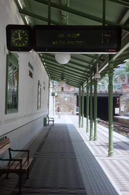 a train station with a bench, a clock, and a ticker reading, we ride with pride.