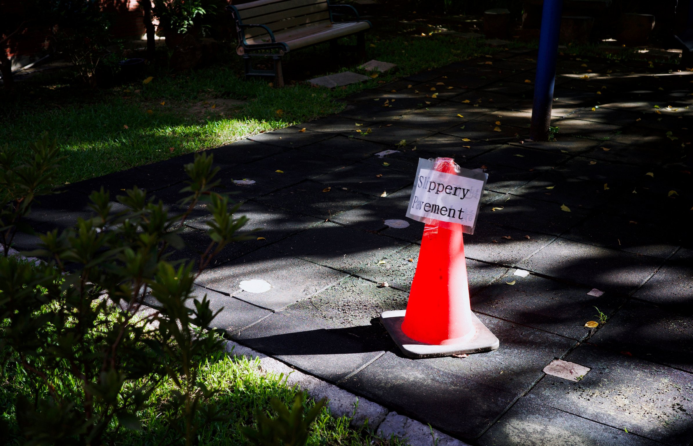 A piece of paper with the text “Slippery Pavement” attached to a traffic cone on the edge of a tiny playground on a sunny day. There’s also an empty park bench.