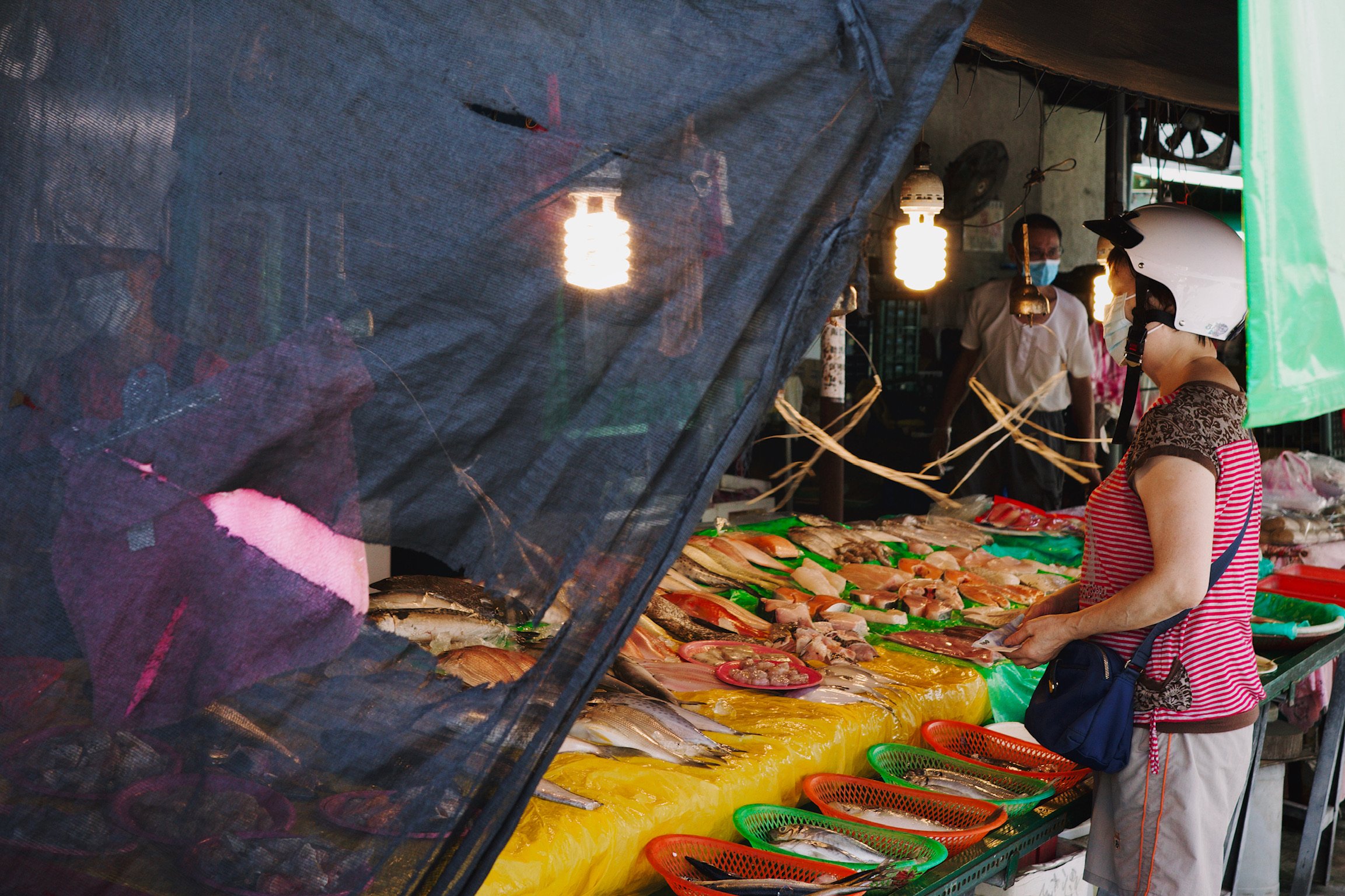 A woman wearing a scooter helmet buying fish from a vendor with bright yellow and green tarp.