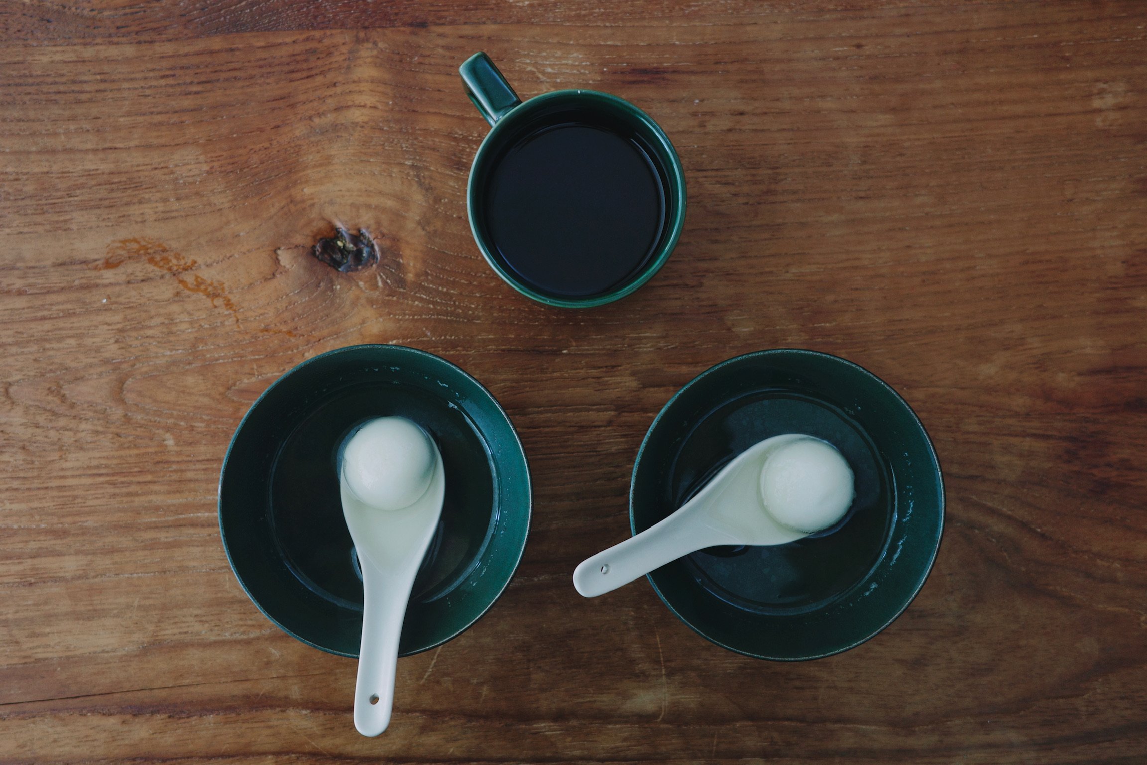 Two green bowls each with one sweet rice dumpling in a white spoon, and a green mug with black coffee. All on a wooden table.