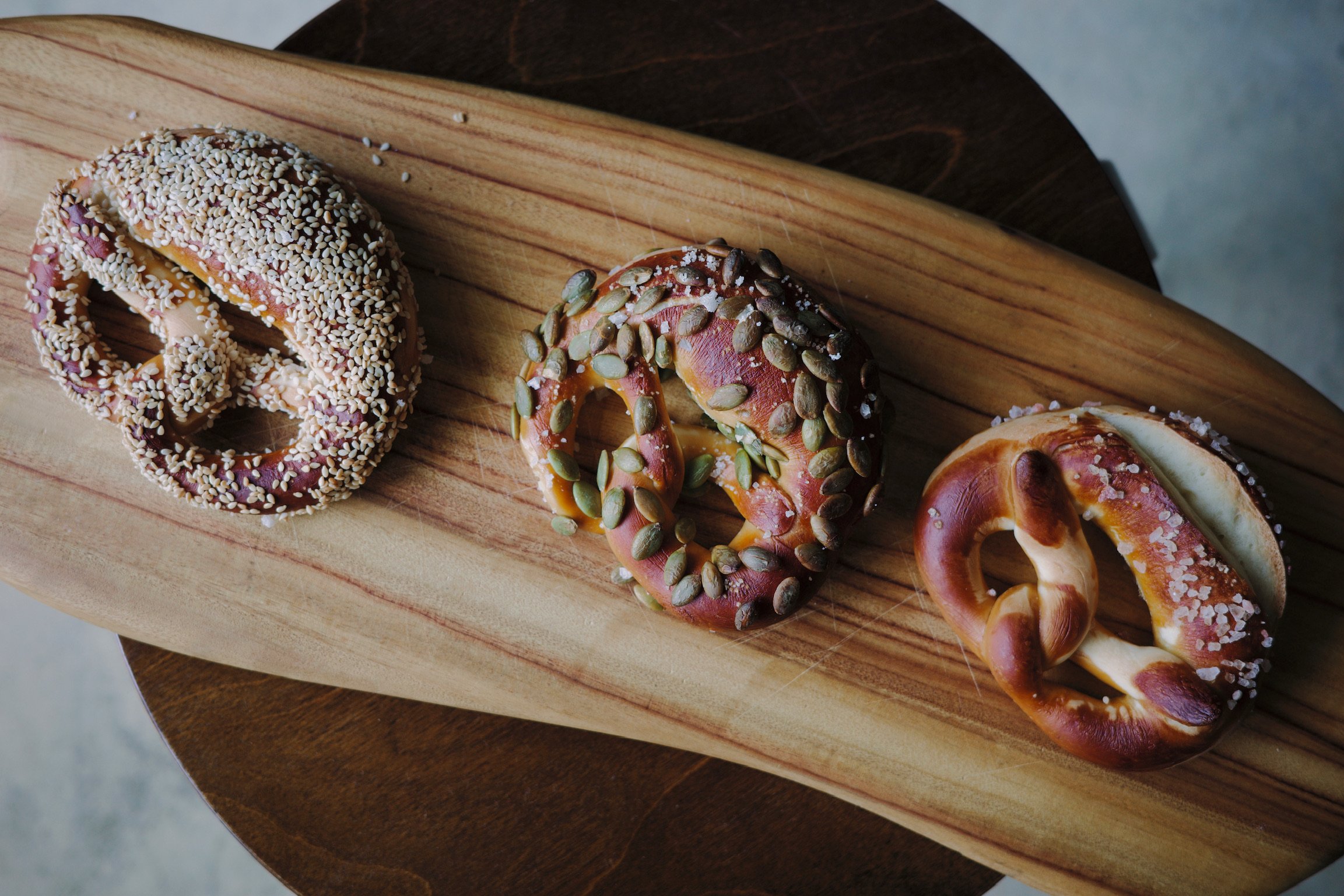 Overhead shot of three pretzels, one covered in sesame, one covered in pumpkin seeds, and one lightly salted, in a row on a irregularly shaped wooden board, on top of a dark walnut stool.