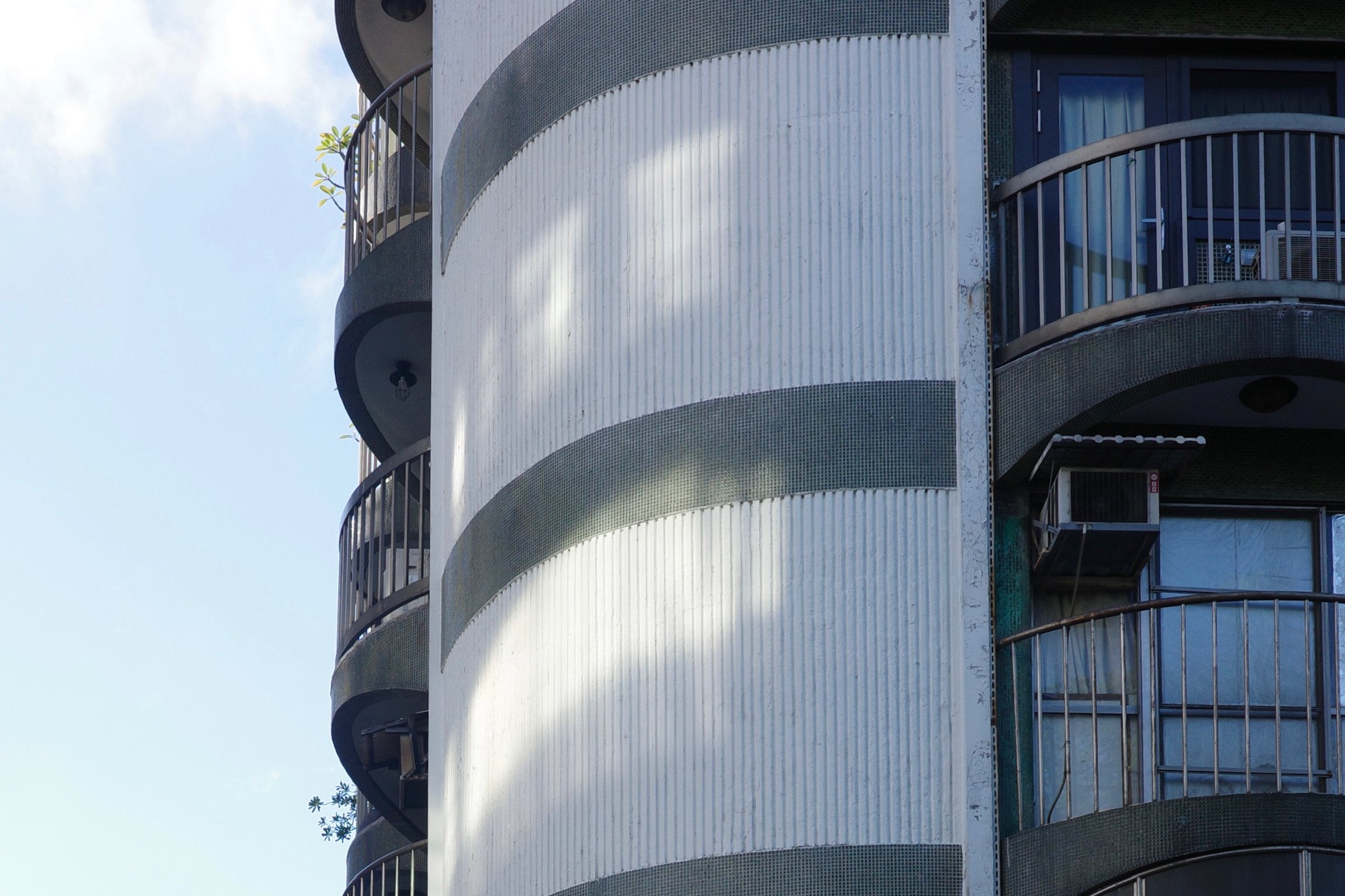 Shadow casted on a white tiled building with faded dark green strips and curved corners. Semicircle-shaped balconies. Clear blue sky.