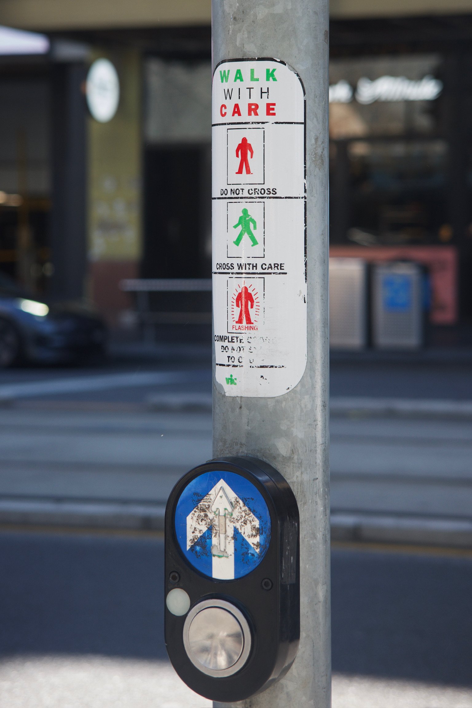 a sign on a pole for people to hit the button for crossing. red figure on the top, green figure in the middle walking, and a red muscle-y figure with shiny symbols around them at the bottom.