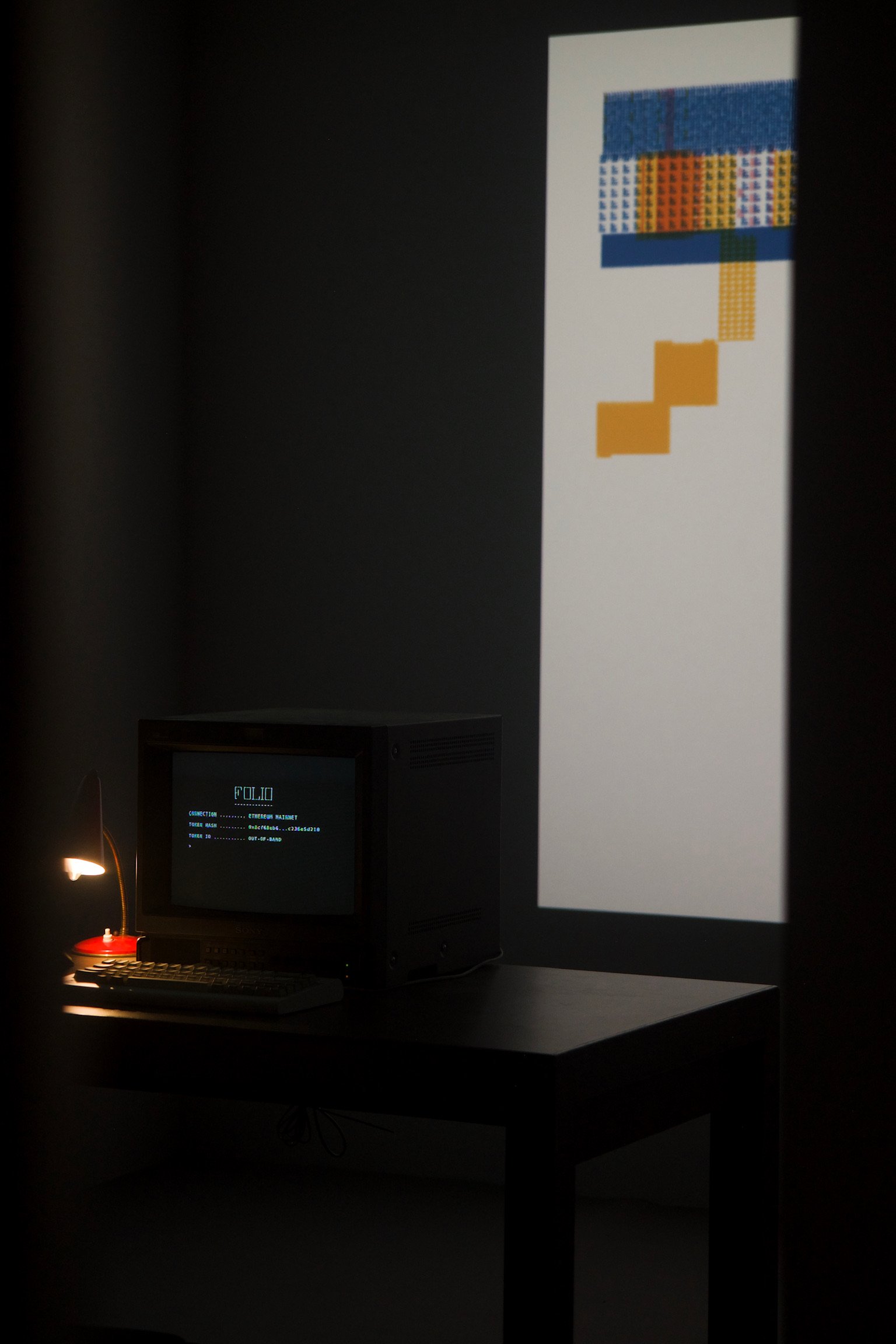 An image projected on a wall. In front of the wall a table in the middle of a dimly lit room. On the table, an old school computer monitor showing a terminal, a mechanical keyboard, and a lamp.