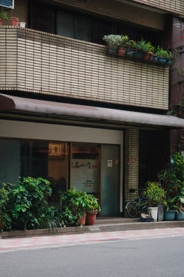 street shot, asphalt, shopfront with lots of potted plants, a row of color plant pots on the second floor, finely tiled beige building.
