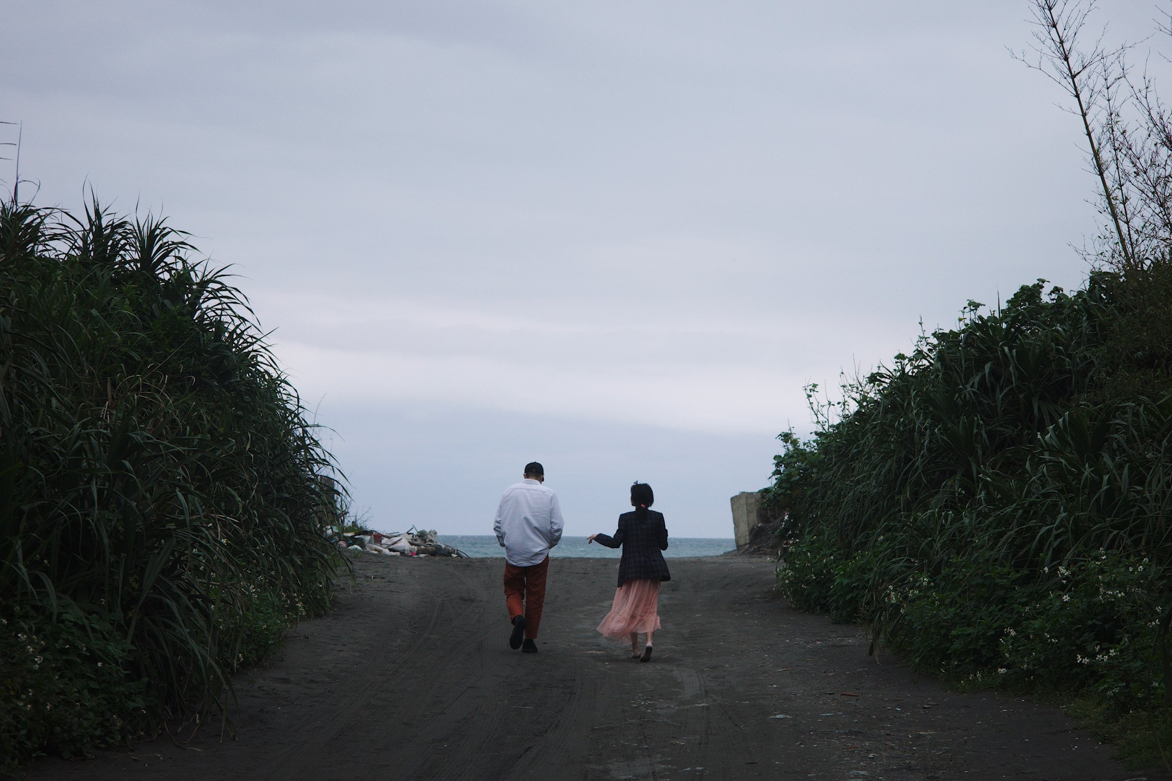 A man and a woman walking towards a beach on a cloudy day.