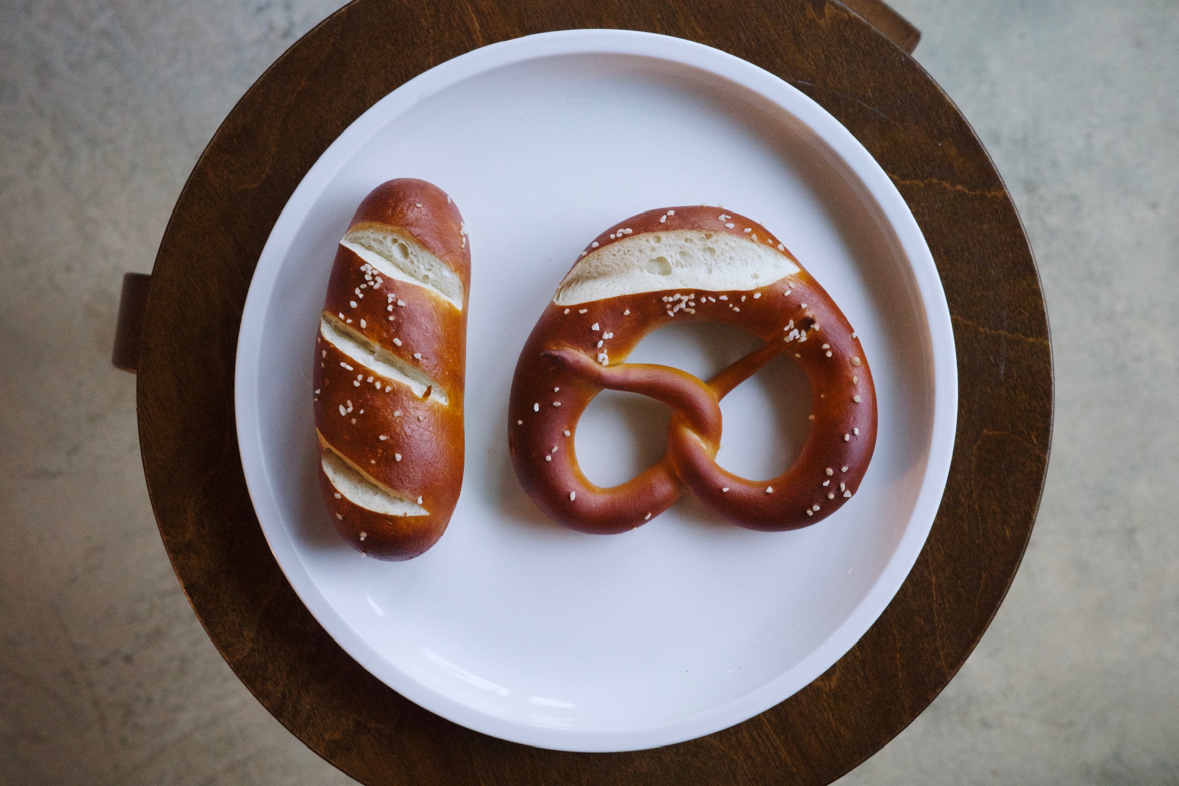 One pretzel and one pretzel stick side by side on a white plate on a stool.