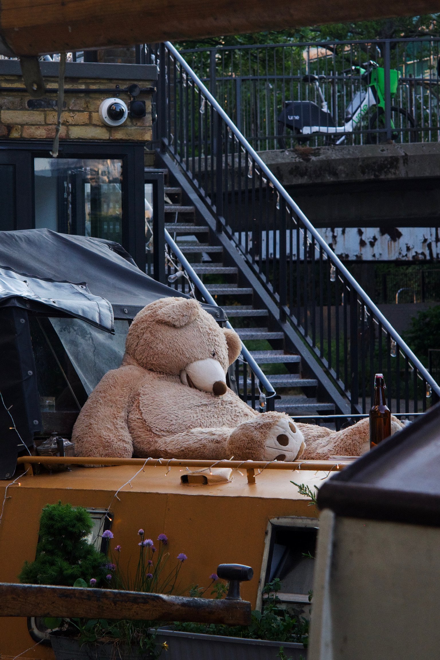A giant bear plushy looking defeated on the roof of a house boat with a beer bottle by it.