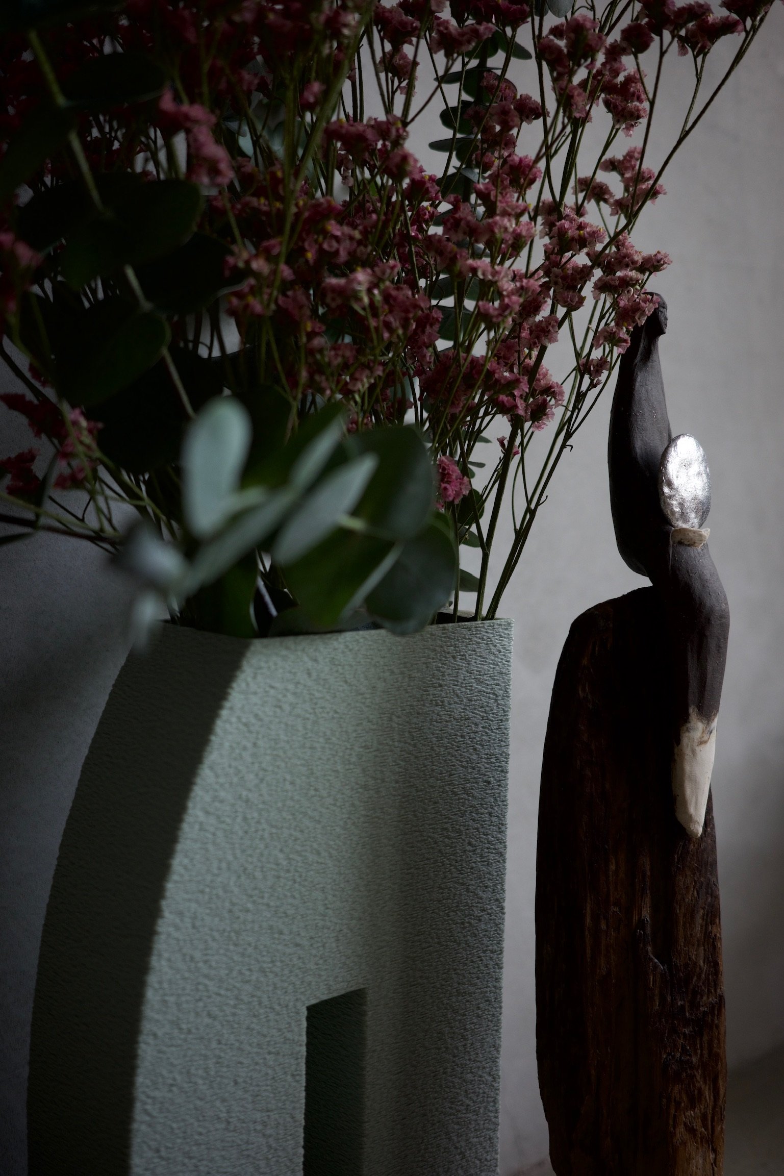 A sculpture of a woman sitting on a block of wood holding a silver disc. Behind her, a textured 3D printed curved vase with flowers and eucalyptus.