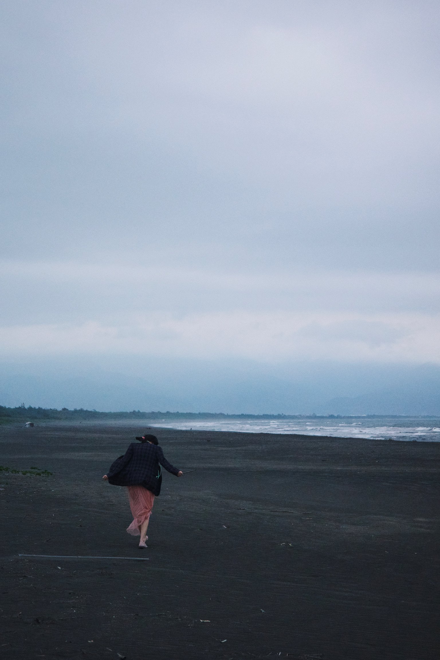 Woman wearing a pink dress and a blazer running on a gray beach on a cloudy day.