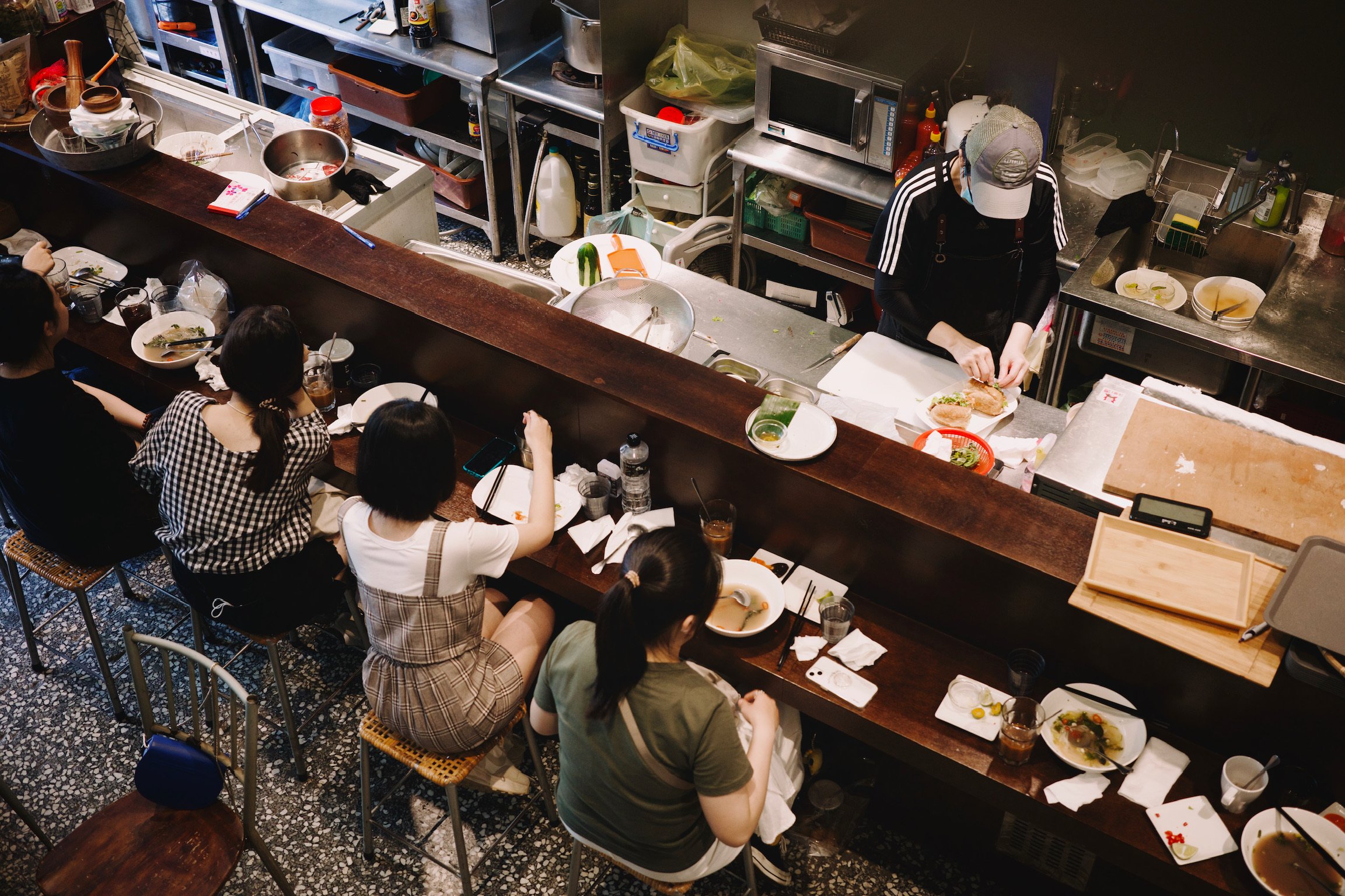 Top down shot of a restaurant bar counter with a single staff working on food, and four women customers sitting in front of finished bowls of phos.