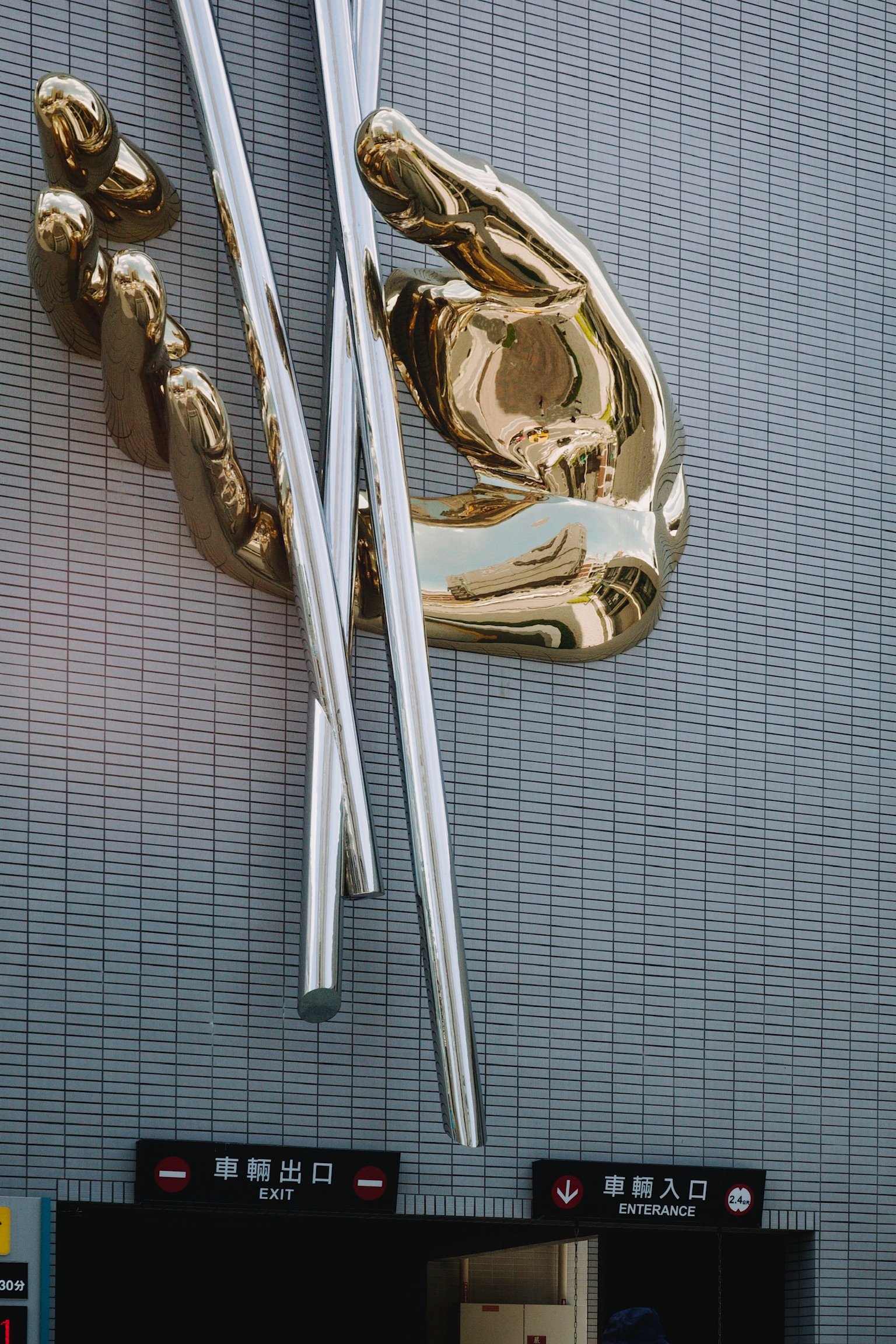 A sculpture, of giant golden hand holding what looks like aluminum rods or straws floating in space, on the side of a finely tiled building in white.