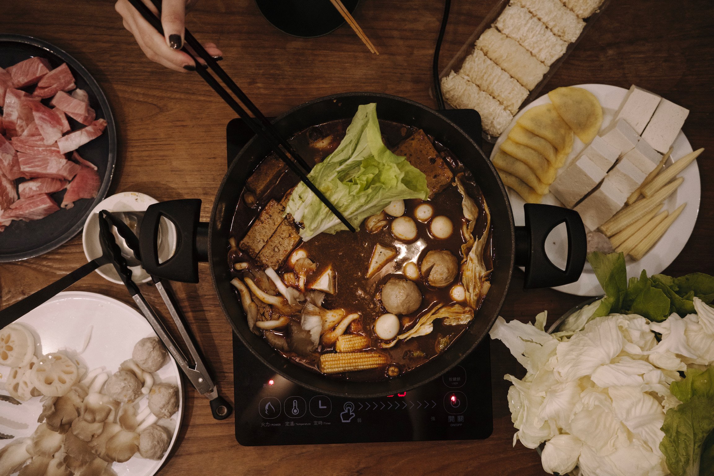 Top down shot of a hot pot with platters of vegetable and meat and one single woman’s hand holding a pair of chopsticks.