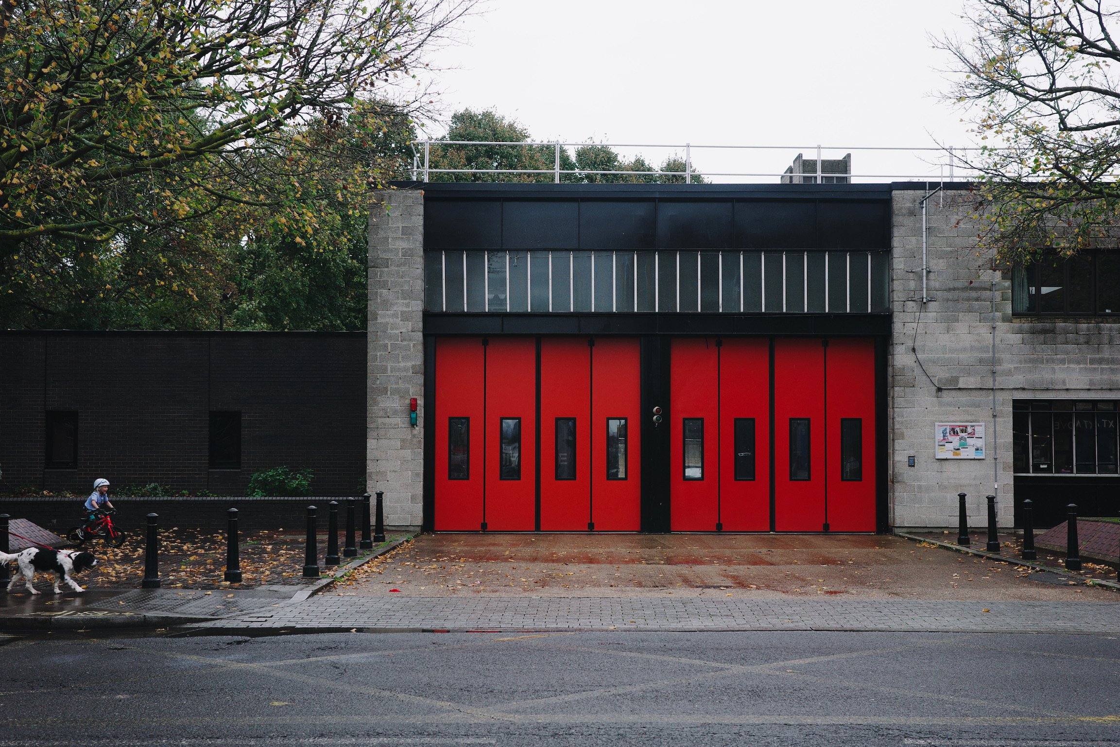 A brutalist fire station with read doors and exposed concrete. A dog and a child appears on the edge of the photo.