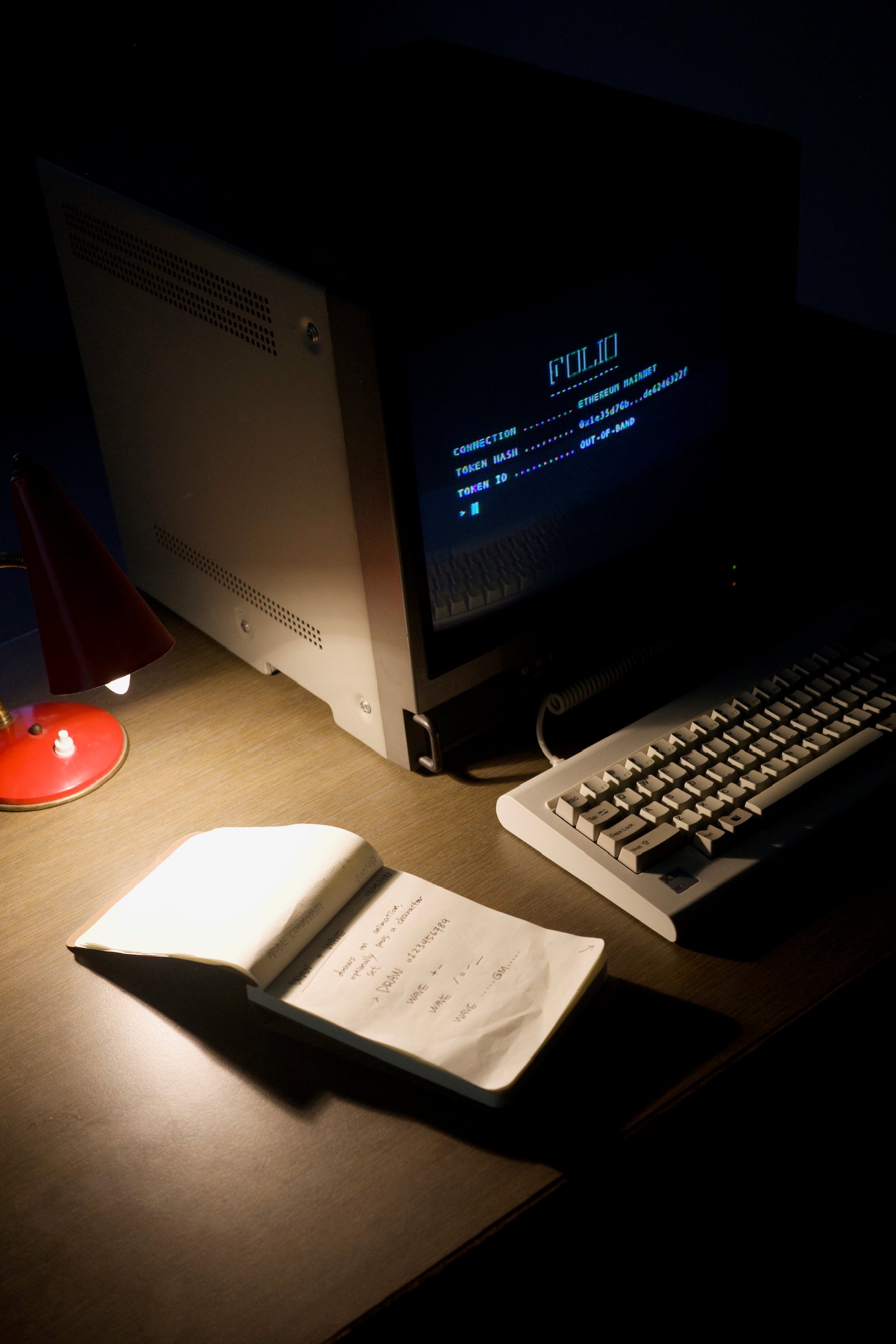 An old school computer monitor showing a terminal, a mechanical keyboard, a lamp, and a notebook listing commands, in a dimly lit room.
