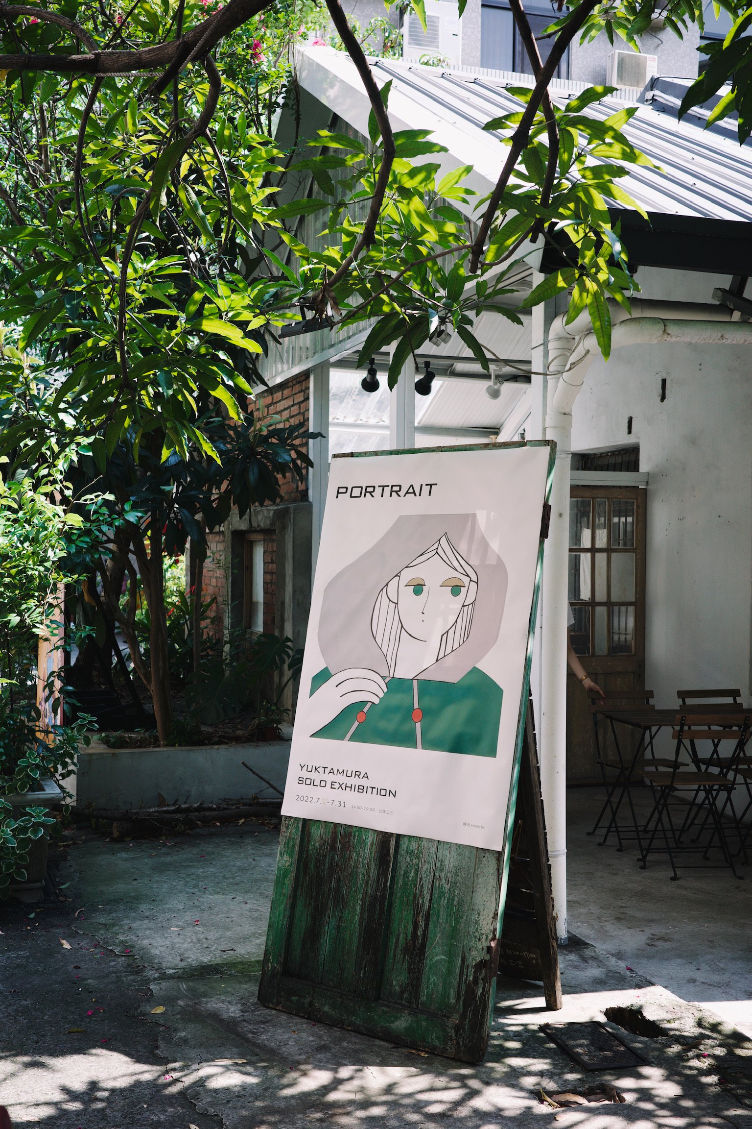 An aged dark green wooden panel, with a white poster reading Portrait, Yuktamura Solo Exhibition, stood in the front yard of a small, old, white structure.