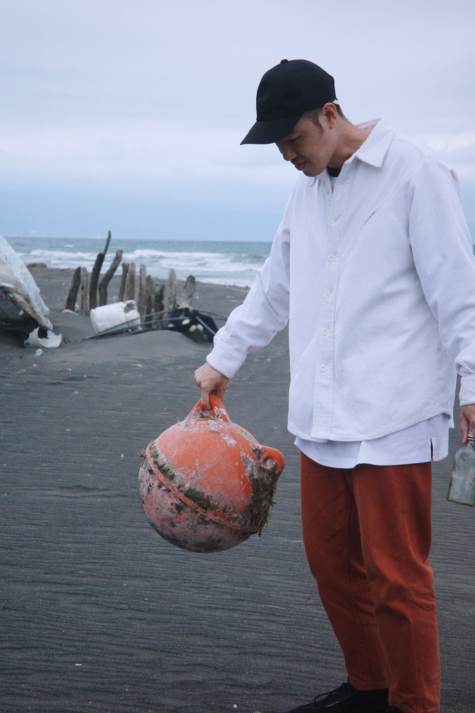 Man wearing a black cap, white shirt, orange pants, black shoes, holding a pinkish orange buoy on a beach with ocean in the background.