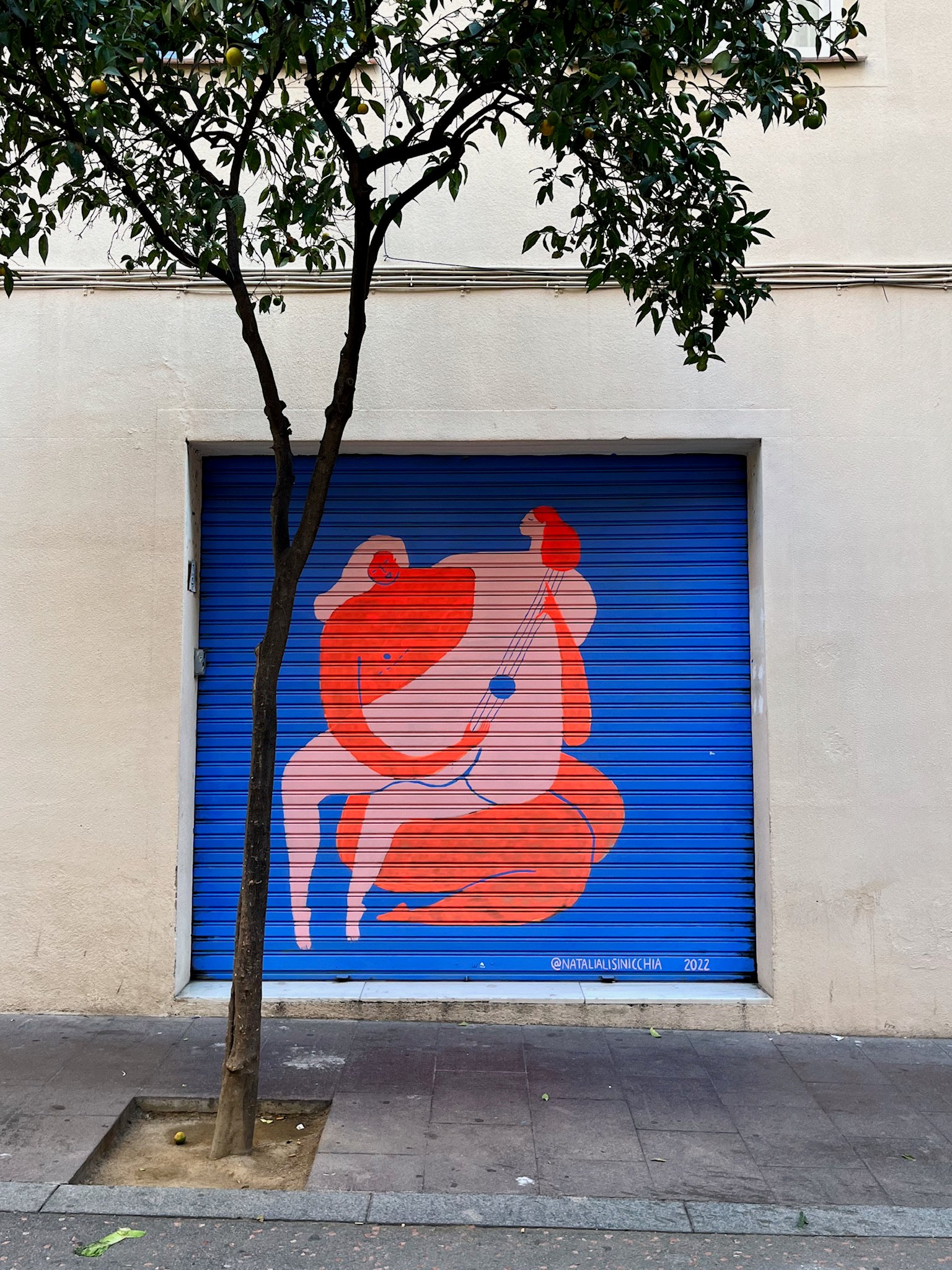 Barcelona street art with bright blue background and a woman sitting on another woman with her back shaped like a string instrument, being played.
